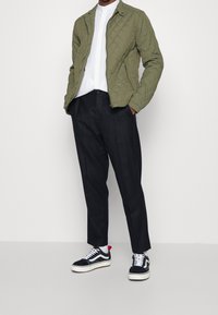 Grüne gesteppte Jacke mit Reißverschluss und Kragen, weißes Shirt, schwarze maßgeschneiderte Hose und schwarze Sneakers mit weißen Akzenten und roten Details.