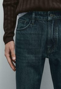 Primo piano di una persona che indossa jeans in denim blu scuro e un maglione marrone a coste, con una mano rilassata accanto al corpo.