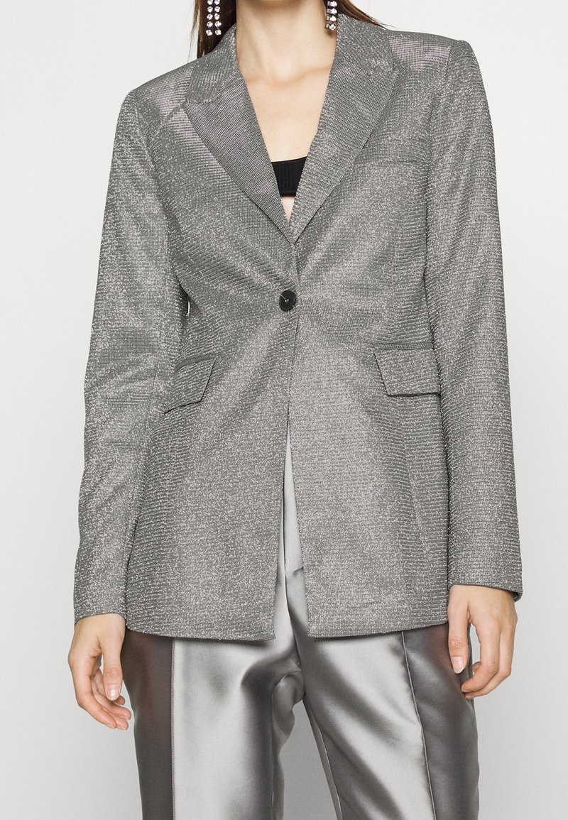 Femme portant un blazer argenté scintillant à bouton unique avec poches à rabat, associé à un pantalon bicolore argenté brillant et des boucles d'oreilles pendantes.