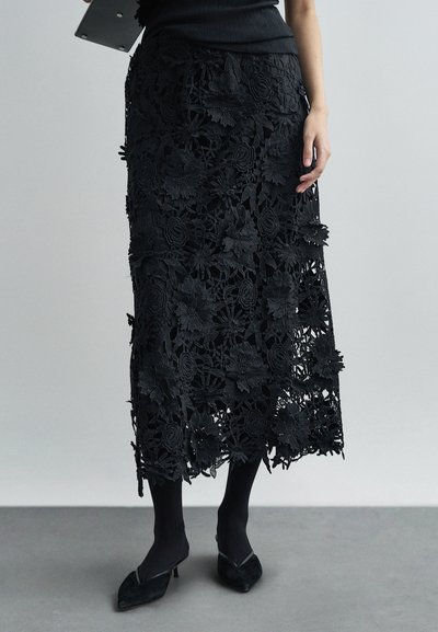 Falda de encaje negra con diseño floral, que presenta detalles recortados y un dobladillo con volante. Llevada con medias negras y tacones de punta.