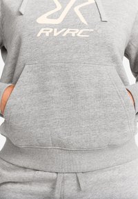 Grå hoodie med frontficka, händer i fickorna, och vit "RVRC" logotyp på bröstet, matchad med grå sweatpants.