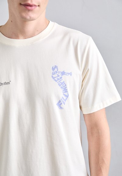 T-shirt en coton couleur crème avec un design graphique bleu portant le texte « rythme » et une silhouette de figure humaine sur le devant.