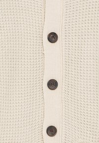 Krämfärgad stickad kofta med ett strukturerat mönster, fem mörkbruna knappar och ribbade kanter längs knappfliken.