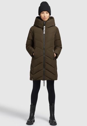 Olijfgroene pufferjas met een hoge kraag, frontale rits en chevronstiksel. Gecombineerd met zwarte leggings en stevige zwarte laarzen. Zwarte gebreide muts.