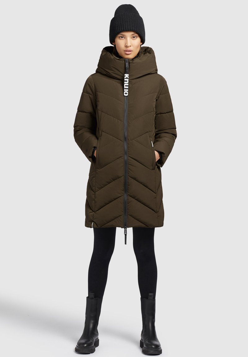 Oliven Grønne pufferjakke med høj krave, frontlynlås og chevron-søm. Kombineret med sorte leggings og kraftige sorte støvler. Sort strikket hat.