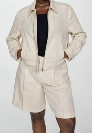 Osoba ubrana w beżową kurtkę zapinaną na zamek i dopasowane beżowe spodenki, na czarnej bluzce, stojąca z rękami w kieszeniach kurtki.