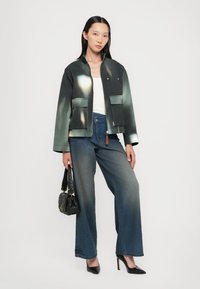 Schwarze und grüne Gradient-Jacke mit zwei Vordertaschen, kombiniert mit weit geschnittenen Denim-Jeans. Das Model hält eine kleine schwarze Tasche und trägt High Heels.