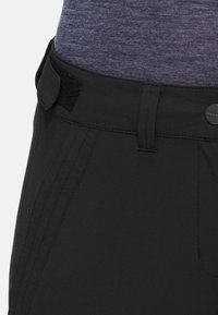 Pantalon noir à taille avec sangle latérale réglable en Velcro, passants de ceinture et détail de bouton à l'avant, porté avec un haut bleu foncé chiné.