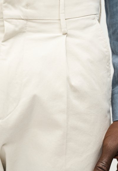 Gros plan sur un pantalon beige avec des plis, une passoire de ceinture et une main brune reposant sur le côté.