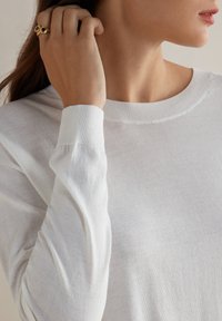 Biały, lekki sweter z prostym okrągłym dekoltem, prążkowanymi mankietami i gładką fakturą. Do ważniejszych detali należy subtelny połysk.