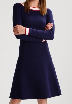 Femme portant une robe bleu marine à manches longues avec des rayures blanches et rouges sur l'encolure et les poignets, les bras croisés sur la poitrine.