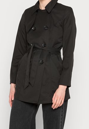 Femme portant un manteau noir à double boutonnage avec des boutons et une ceinture nouée, associé à un pantalon gris foncé sur un fond uni.