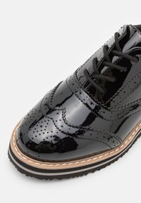 Fekete patent bőr öltönycipő díszes brogue részletezéssel, kerek orral, fekete fűzőkkel, és kontrasztos bézs és fekete talppal.