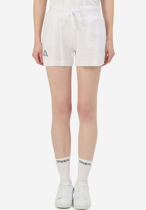 Persona che indossa pantaloncini bianchi con coulisse e tasche frontali, calzini bianchi con logo Kappa e sneakers bianche, in piedi davanti a uno sfondo semplice.