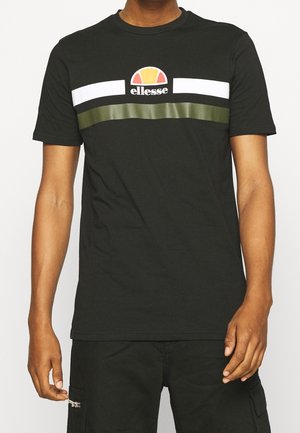 Mężczyzna ubrany w czarną koszulkę ellesse z białymi i oliwkowozielonymi poziomymi paskami na wysokości klatki piersiowej, w połączeniu z czarnymi spodniami z kieszenią na zamek.