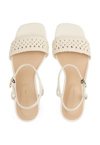 Sandales blanches avec une bride tressée, bout carré et bride à la cheville. Texture en cuir lisse, avec une semelle intérieure beige et des détails en métal doré.