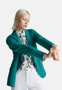 Blondýnka natahující ruce dopředu, oblečená v tyrkysové saku, bílé květinové košili na knoflíky a bílých kalhotách na jednobarevném pozadí.