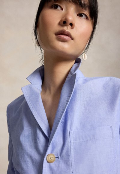 Νέα γυναίκα που φοράει ένα ανοιχτό μπλε πουκάμισο με κουμπιά και γιακά ανασηκωμένο, με σκουλαρίκι κοχύλι, κοιτάζοντας ελαφρώς προς τα πάνω και πλάγια.