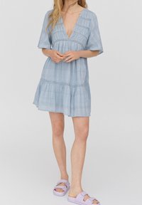 Světle modré šaty po kolena s hlubokým véčkovým výstřihem, krátkými rukávy, zřasenou texturou a vrstevnatým lemem. Doplnené lila nazouvacími sandály.