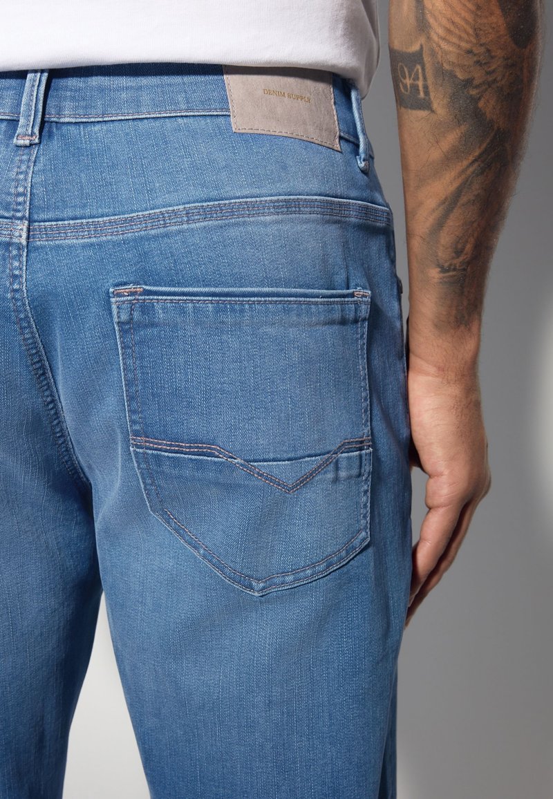 Gros plan d'une personne portant un jean en denim bleu clair avec une poche arrière visible et un bras tatoué.