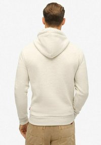 Jasne beżowe hoodie z kapturem na sznurku, wykonane z miękkiej tkaniny. Posiada ściągacz u dołu i na mankietach, z małą metką logo z boku.