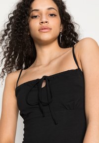 Top preto sem mangas com decote franzido e detalhe de laço ajustável na frente; tecido liso com forma ajustada e acentos mínimos.