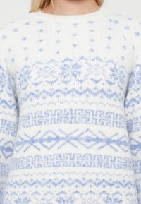 Λευκό πλεκτό πουλόβερ με ανοιχτό μπλε νορβηγικού στυλ σχέδια που περιλαμβάνουν νιφάδες χιονιού και γεωμετρικά σχήματα, το οποίο φοράει ένα άτομο με ξανθά μαλλιά.
