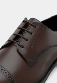 Καφέ δερμάτινο παπούτσι με κορδόνια, με λείο φινίρισμα, διάτρητο κάλυμμα μύτης και διακριτικές ραφές. Σκούρα κορδόνια συμπληρώνουν το σχέδιο.