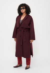 Burgundy long coat with wide collar, button closure, waist tie, and side slits. Worn over gray top and paired with red pants and pointed shoes.