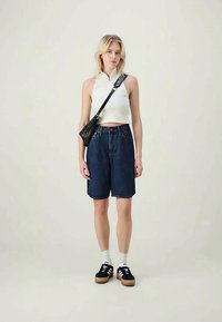 Blonde woman wearing sleeveless white cropped top, dark denim knee-length shorts, black crossbody bag, white socks, and black sneakers.