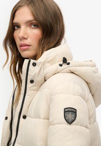 Beżowa kurtka puchowa z kapturem, z czarnym zamkiem błyskawicznym i guzikami. Na lewym rękawie znajduje się czarna łatka z logo płatka śniegu. Miękka w dotyku.