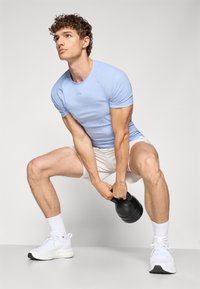 Lys blå treningstrøye og hvite shorts brukt av en person som gjennomfører en knebøy, holder en svart kettlebell. Hvite joggesko og ankellange sokker er synlige.