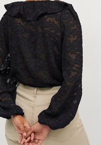 Schwarze Spitzenbluse mit einem Rüschenkragen, langen Puffärmeln und floralen Muster; geschichtete Textur vor einem hellen Hintergrund sichtbar.