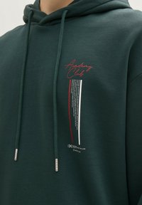 Ciemnozielona bluza z kapturem z nadrukiem na przodzie, na którym znajduje się napis "Academy Club" w kolorach czerwonym, białym i czarnym. Srebrne końcówki na sznurków.