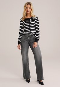 Cardigan rayé noir et blanc avec des boutons ; associé à un jean gris à taille haute et jambes larges et des escarpins noirs à bout pointu.