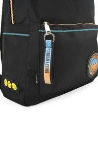 Mochila negra con un acabado suave y texturizado, que presenta un bolsillo delantero con cremallera y costuras azules, parches de cara sonriente y una correa de marca.