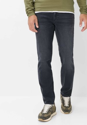 Uomo che indossa jeans scuri, sneakers verdi e bianche e un maglione verde oliva, in piedi su sfondo bianco.