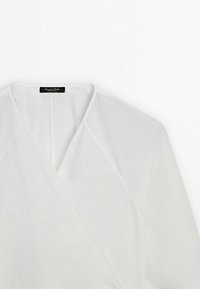 Blouse blanche à enrouler en tissu léger. Possède un décolleté en V et des manches triangulaires avec des lignes épurées et sans quincaillerie ni motifs visibles.