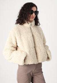 Krémově zbarvená, plyšová fleecová bunda s vysokým límcem, předním zipem a oversized střihem, spárovaná se světle hnědými kalhotami.