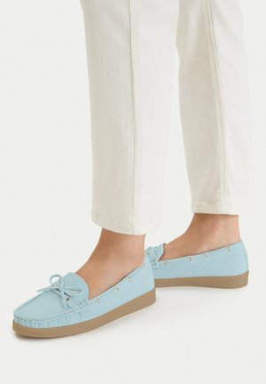 Jasnoniebieskie wsuwane mokasyny z kokardą, noszone z beżowymi spodniami, pokazane z boku na jednolitym jasnym tle.