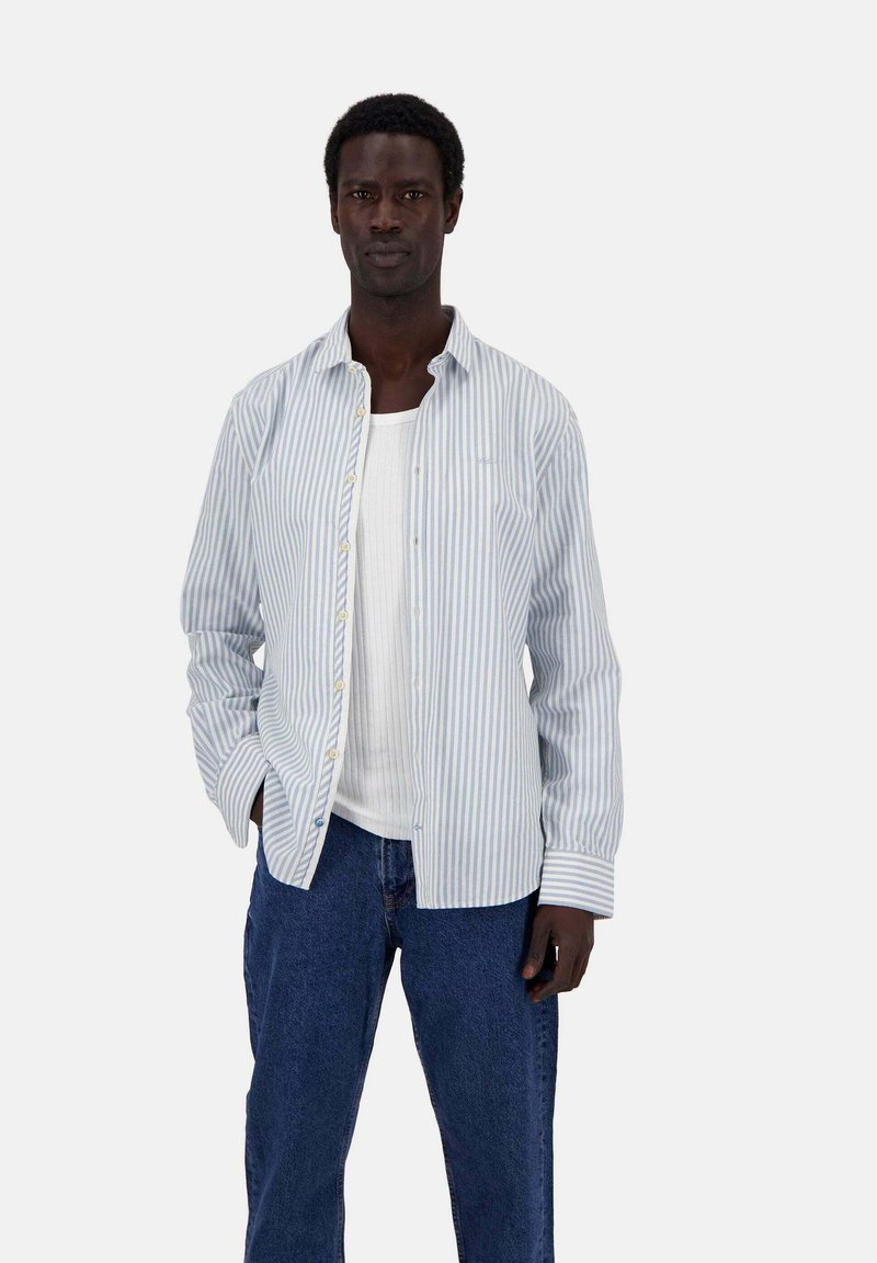 Camicia a righe blu chiaro e bianca, con colletto e maniche lunghe, indossata sopra una canotta bianca, abbinata a jeans blu.