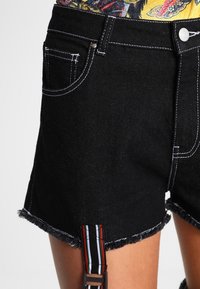 Svarta denimshorts med fransad nederkant, vit söm och sidoflik med röda, gröna och blå ränder. Två framfickor synliga.