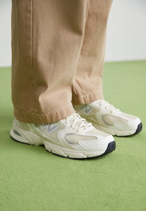 Beżowe spodnie zakrywające kostki oraz białe buty sportowe New Balance stojące na jasnozielonej, wykładzinowej podłodze.