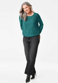 Blusa lungo maniche color teal abbinata a jeans dritti grigio scuro. Tacchi alti neri lucidi completano l'outfit. Texture del tessuto liscia.