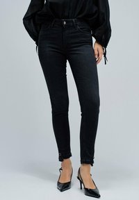 Jeans skinny negros con una textura de denim suave, tiro alto y botones en la parte frontal; el dobladillo presenta bucles únicos para un ajuste ajustable.