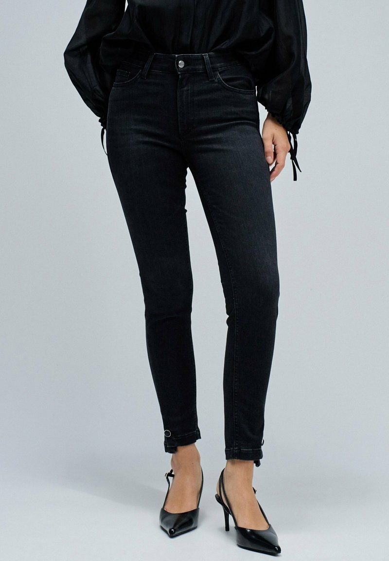 Jeans skinny negros con una textura de denim suave, tiro alto y botones en la parte frontal; el dobladillo presenta bucles únicos para un ajuste ajustable.