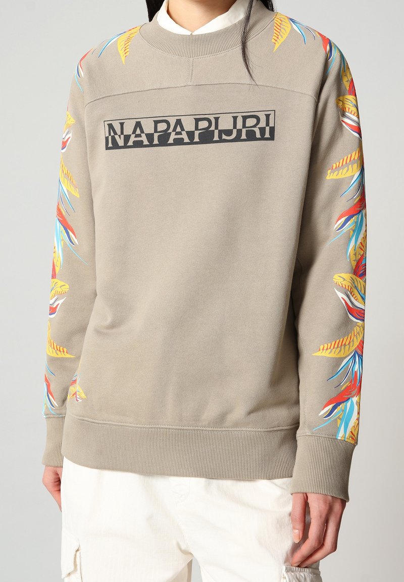 Sweat-shirt beige avec des motifs colorés de feuilles tropicales sur les manches et le logo "NAPAPIJRI" sur la poitrine, porté par-dessus une chemise blanche.