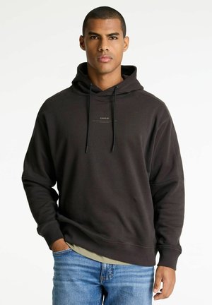 Mann in schwarzer Kapuzenjacke mit dem Text "CHASIN'", hellgrünem Unterhemd und blauer Jeans, steht mit einer Hand in der Tasche vor weißem Hintergrund.