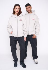 Svetlošedé mikiny na zips s vreckami a vyšitým logom, nosené s čiernymi cargo nohavicami a teniskami, predvádzané dvoma modelmi.