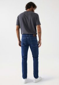 Camiseta de color gris oscuro y jeans azules de corte recto. Los jeans tienen dos bolsillos traseros, un parche de cuero marrón y zapatillas de deporte blancas.